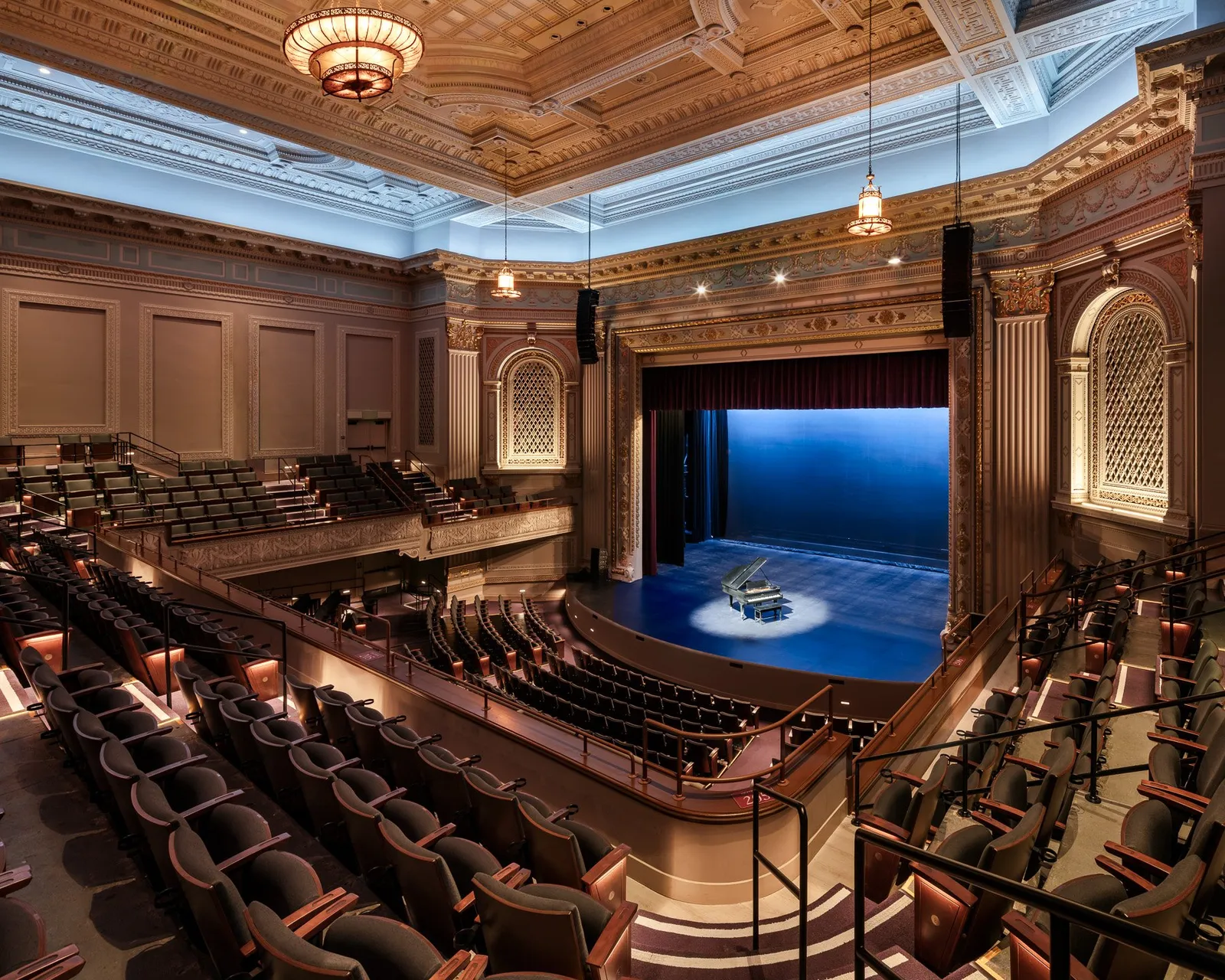 The restored Calvin Simmons Theater from high at the side: a coffered ceiling and chandelier above curved rows of seats, a piano lit on a blue stage.